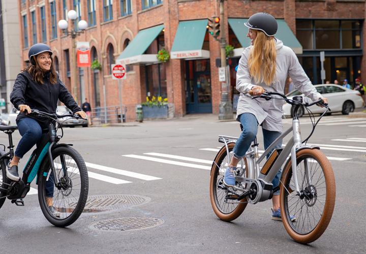 Two women getting ready to start pedaling their electric bikes on a busy city street.